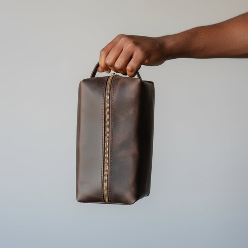 Brown leather toiletry bag held by a hand against a plain background