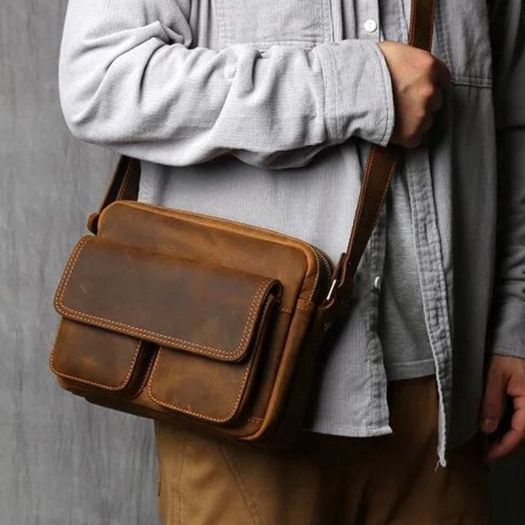 Brown leather bag carried by a person wearing a gray shirt against a neutral background