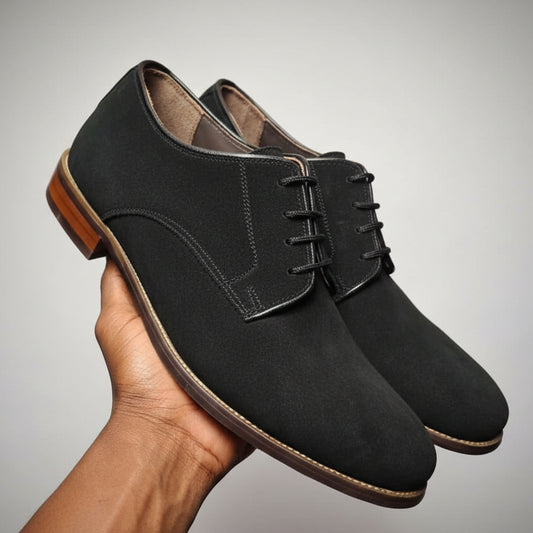 black suede shoes held in a hand with a wooden floor and shoes in the background