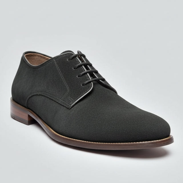 Pair of black suede shoes held by a hand with a wooden floor and shoes in the background.